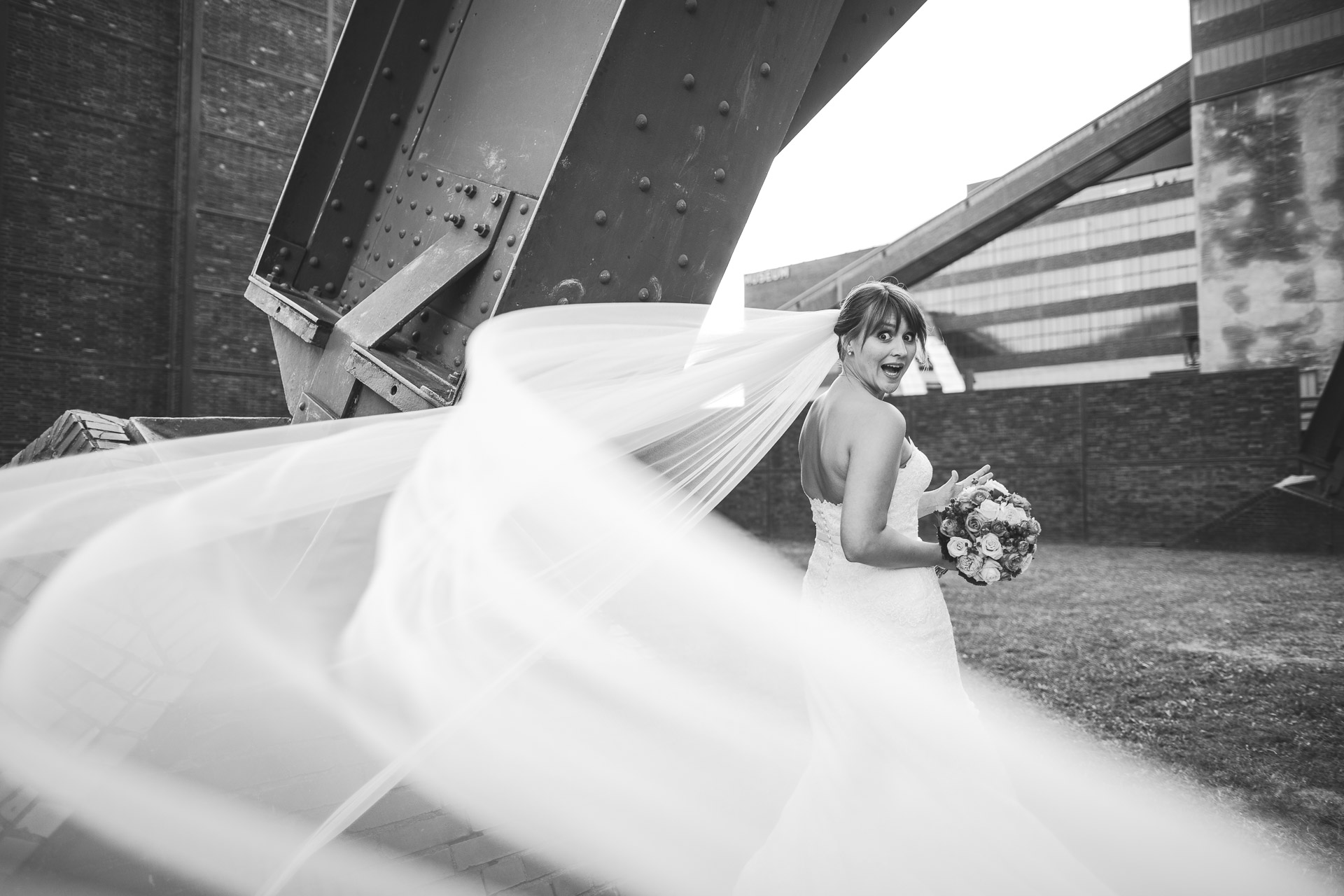 Witziges Hochzeitsfoto mit Braut auf Industriegelände. Schleier fliegt weg.